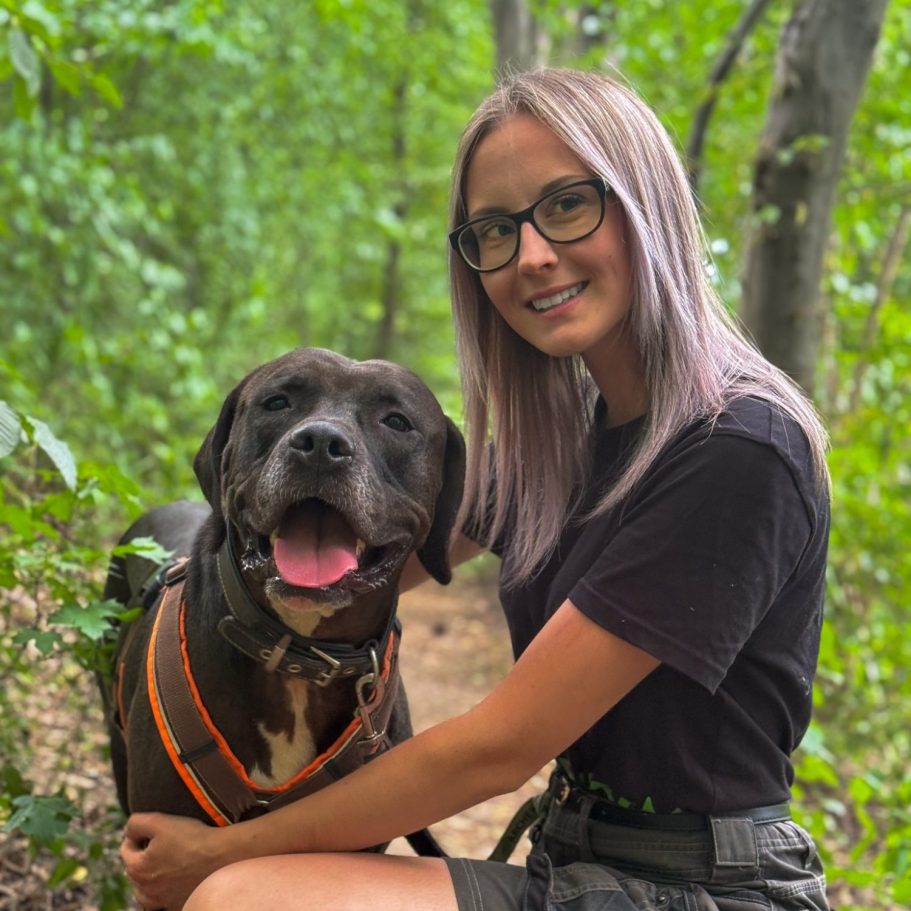 Junge Tierpflegerin sitzt neben einem Tierheimhund auf einem Waldweg umgeben von Bäumen.