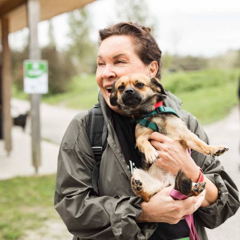 Ehrenamtliche Spaziergeherin der Arche Noah mit einem kleinen Hund in den Armen, lächelt glücklich im Freien.