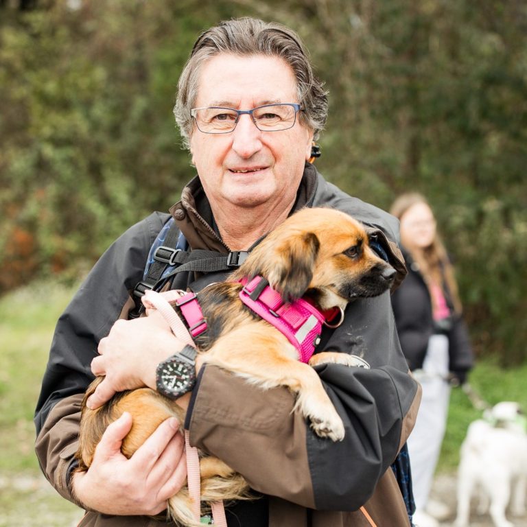 Mann mit Brille hält einen kleinen Tierheim-Hund in den Armen, im Hintergrund eine weitere Person. Tierschutz:läuft2024