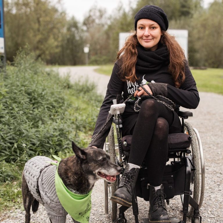 Eine Frau im Rollstuhl mit ihrem Hund, der einen grünes Halstuch trägt, auf einem Weg. Tierschutz:läuft2024