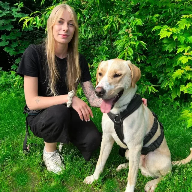 Mann mit kurzen Haaren kniet neben einem Hund im grünen Gras.