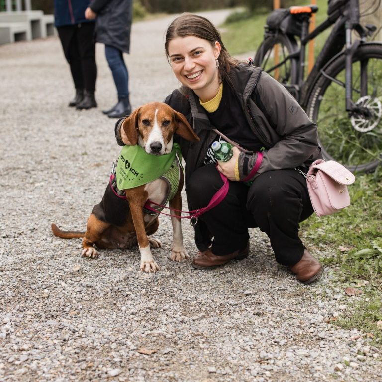 Eine Person sitzt neben einem Tierheim-Hund mit grünem Halstuch auf einem Weg. Tierschutz:läuft2024