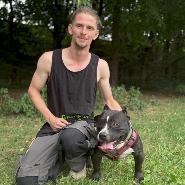 Mann mit kurzen Haaren kniet neben einem Hund im grünen Gras.