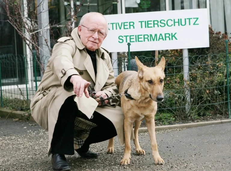 Der Gründer des Vereins, Herbert Oster, in einem Trenchcoat kniet neben einem Hund vor dem Eingang des Tierheims Arche Noah.