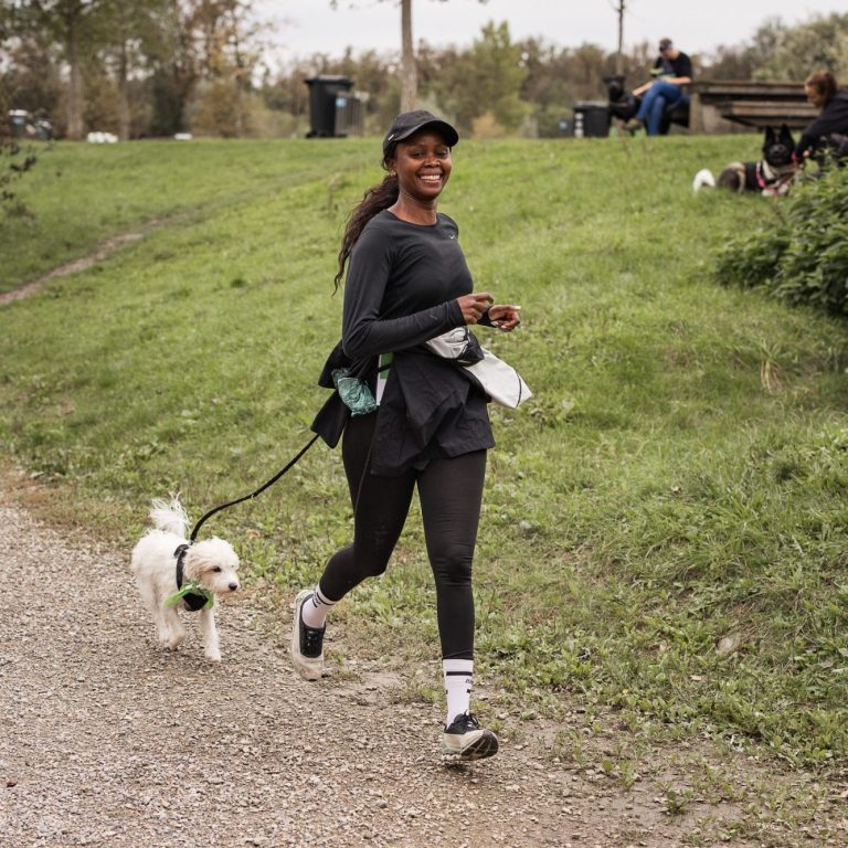 Eine Frau joggt mit ihrem kleinen Hund entlang eines Weges in einem Park. Tierschutz:läuft2024