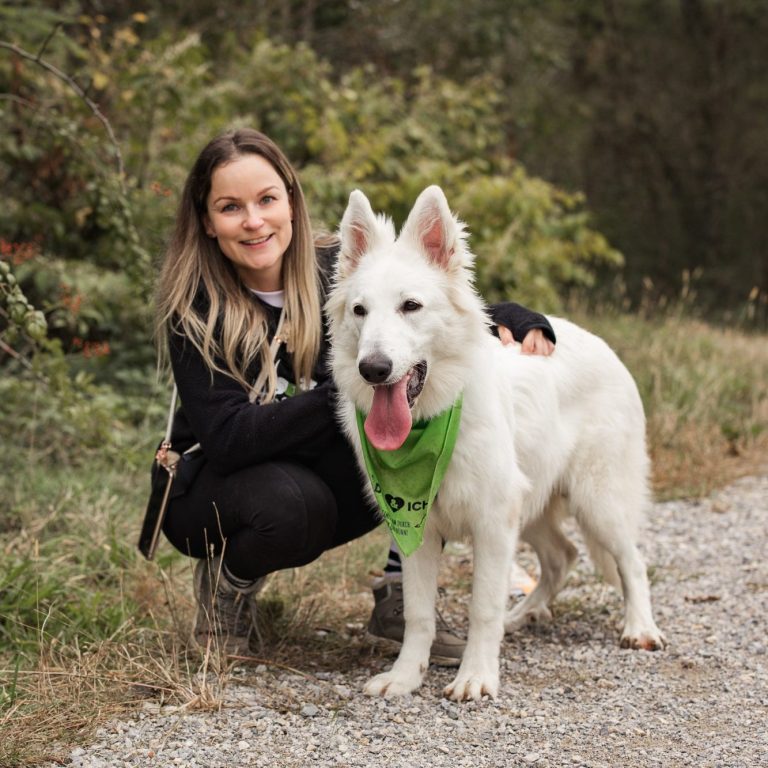 Arche Noah - Aktiver Tierschutz Austria: Ehrenamt Spaziergeherinnen Eine Frau kniet neben einem weißen Hund in der Natur. Der Hund trägt ein grünes Tuch auf dem steht 'du & ich - gemeinsam durch dick & dünn'.