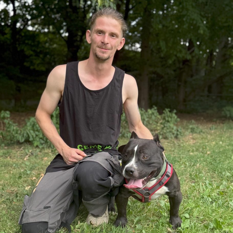 Junger Mann kniet neben einem Hund auf einer Wiese.